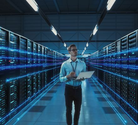 Technician walking through a data center aisle with a laptop between illuminated server racks.